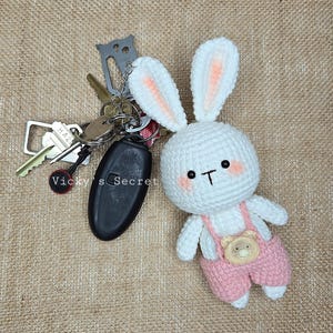 Op de afbeelding: Witte gehaakte konijnen sleutelhanger met roze overall en een bruine berenknop. Het konijn heeft zwarte ogen en een witte neus. De sleutelhanger is bevestigd aan een sleutelbos.