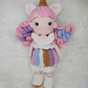Op de afbeelding: Een gehaakte eenhoornpop met roze en paars haar, een witte jurk met kleurrijke strepen en bruine laarzen. De pop draagt een bruine ijshoornhoed.