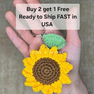Op de afbeelding: Een gehaakte zonnebloem sleutelhanger met een groen blad. De sleutelhanger is bevestigd aan een zilveren sleutelring. De tekst "Buy 2 get 1 Free Ready to Ship FAST in USA" is zichtbaar op de achtergrond.