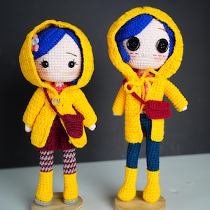May include: Two handmade crochet dolls featuring blue hair, yellow hooded raincoats, and button eyes. The left doll wears a maroon skirt and patterned leggings, while the right doll wears blue pants. Both dolls carry small maroon crossbody bags.