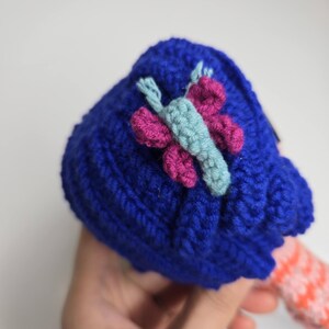 Pu&ograve; includere: Un cappello blu all'uncinetto fatto a mano con un'applicazione a farfalla nei toni del rosa e dell'azzurro. Il cappello &egrave; realizzato con un filato spesso e strutturato. La farfalla &egrave; centrata sul cappello.