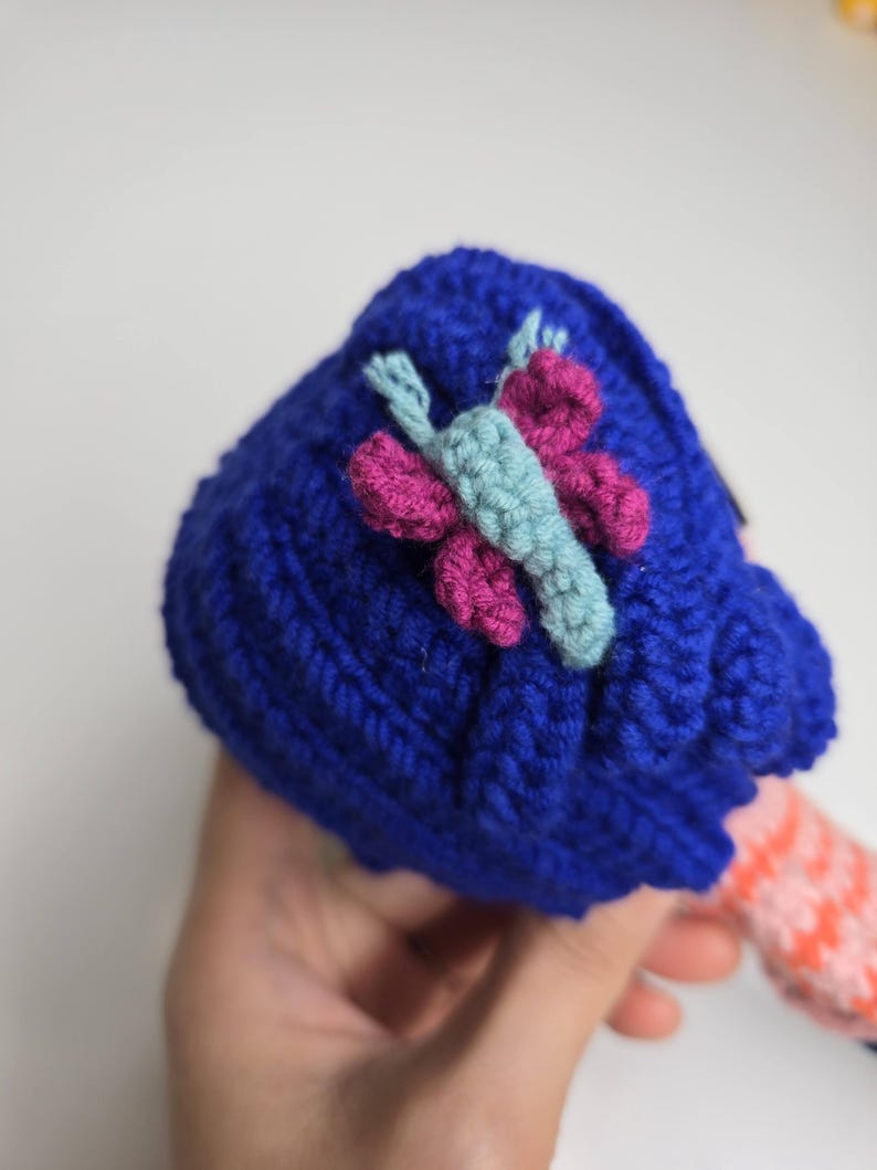 Pu&ograve; includere: Un cappello blu all'uncinetto fatto a mano con un'applicazione a farfalla nei toni del rosa e dell'azzurro. Il cappello &egrave; realizzato con un filato spesso e strutturato. La farfalla &egrave; centrata sul cappello.