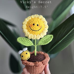 Op de afbeelding: Een gehaakte zonnebloem met een glimlach in een bruine pot met een gehaakte bij. De zonnebloem is geel met een wit centrum en groene bladeren. De pot is bruin met een textuur. De bij is geel met zwarte strepen en witte vleugels. De tekst "Vicky's Secret" en "Height: 7.5" staat boven de zonnebloem.