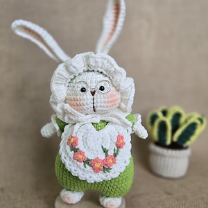 Op de afbeelding: Een gehaakt konijn dat een groene jurk en een witte schort met roze bloemen draagt. Het konijn heeft witte vacht, bruine ogen en draagt ronde brillen. Het konijn staat op een houten voet. De tekst "Vicky's Secret" is zichtbaar onder het konijn.