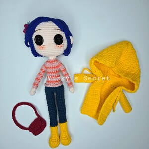 Pu&ograve; includere: Bambola all'uncinetto con capelli blu, una maglia a righe e pantaloni blu scuro, accompagnata da un cappotto con cappuccio giallo e una borsetta rossa. La bambola ha grandi occhi neri a bottone e stivali gialli. Il testo "Vicky's Secret" &egrave; visibile.