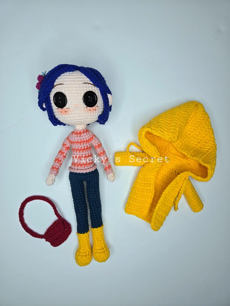 Pu&ograve; includere: Bambola all'uncinetto con capelli blu, una maglia a righe e pantaloni blu scuro, accompagnata da un cappotto con cappuccio giallo e una borsetta rossa. La bambola ha grandi occhi neri a bottone e stivali gialli. Il testo "Vicky's Secret" &egrave; visibile.