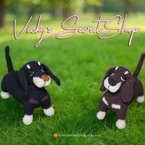 Op de afbeelding: Twee gehaakte teckel hondjes, één zwart en één bruin, staan op groen gras. De honden hebben witte accenten op hun poten, gezichten en ruggen. De shopnaam "Vicky's Secret Shop" staat in sierlijke letters erboven.
