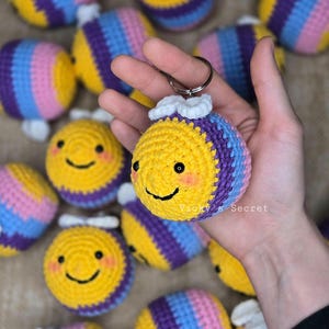 Op de afbeelding: Een verzameling gehaakte bij-versieringen met gele en paarse strepen, witte vleugels en zwarte ogen. De bijen hebben een glimlachend gezicht en houden een sleutelhanger vast.