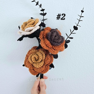 Op de afbeelding: Een boeket van drie gehaakte rozen in tinten bruin, oranje en wit. De rozen zijn bevestigd aan zwarte stelen met zwarte bladeren. Het boeket is gemarkeerd met #2.
