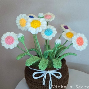 Op de afbeelding: Een gehaakt bloemstuk in een bruine pot met een witte strik. De bloemen zijn wit met gekleurde centra en groene bladeren.