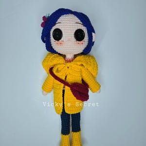 Pu&ograve; includere: Bambola all'uncinetto raffigurante un personaggio con capelli blu, grandi occhi neri a bottone e guance rosee. La bambola indossa un cappotto giallo, pantaloni blu scuro e stivali gialli. Una piccola borsa rossa &egrave; appesa alla spalla. Il testo "Vicky's Secret" &egrave; visibile.