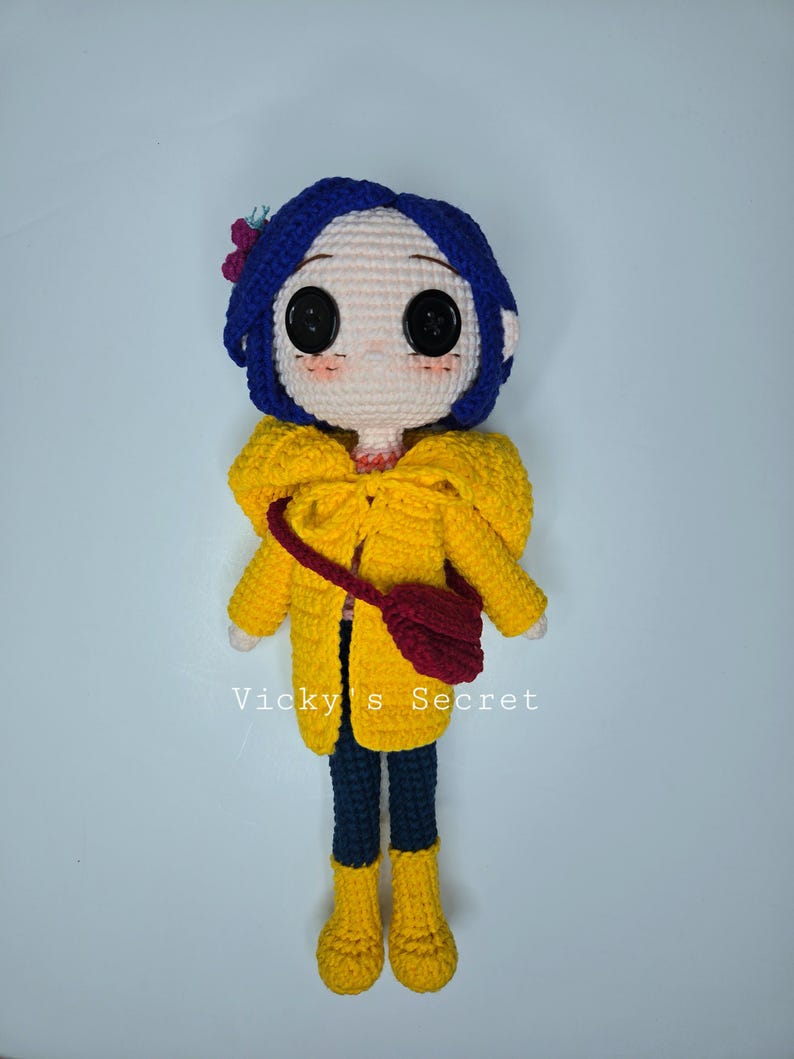 Pu&ograve; includere: Bambola all'uncinetto raffigurante un personaggio con capelli blu, grandi occhi neri a bottone e guance rosee. La bambola indossa un cappotto giallo, pantaloni blu scuro e stivali gialli. Una piccola borsa rossa &egrave; appesa alla spalla. Il testo "Vicky's Secret" &egrave; visibile.