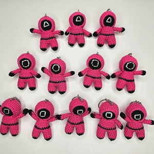 Op de afbeelding: Een verzameling van 10 roze gehaakte figuren met zwarte details. Elk figuur heeft een andere vorm op zijn hoofd, waaronder een driehoek, een cirkel en een vierkant. De figuren dragen allemaal een roze jumpsuit met zwarte strepen.