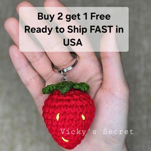 Op de afbeelding: Een rode gehaakte aardbeien sleutelhanger met een groene steel en gele zaden. De sleutelhanger is bevestigd aan een zilveren sleutelring. De tekst "Vicky's Secret" is zichtbaar in de afbeelding.