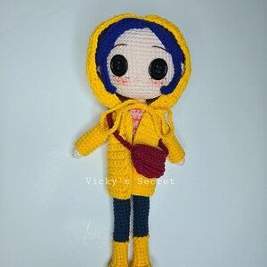 Pu&ograve; includere: Bambola all'uncinetto fatta a mano con impermeabile giallo con cappuccio, capelli blu e una borsa rossa. La bambola ha grandi occhi neri a bottone e indossa pantaloni blu scuro e stivali gialli. Il testo "Vicky's Secret" &egrave; visibile.