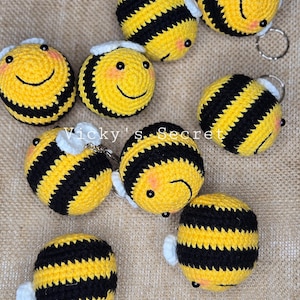Op de afbeelding: Een verzameling handgemaakte gehaakte bijen sleutelhangers. Elke bij heeft een gele body met zwarte strepen, kleine zwarte ogen en een lachend gezicht. De bijen hebben kleine witte vleugels en een zilveren sleutelring. De tekst "Vicky's Secret" is zichtbaar.