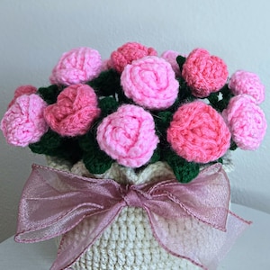 Op de afbeelding: Een gehaakt boeket roze rozen in een crèmekleurige gehaakte pot met een roze tule strik.