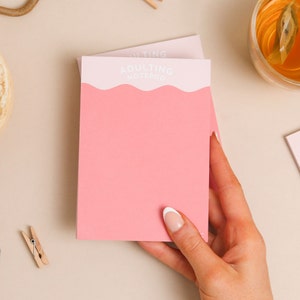 May include: Pink notepad with the text "Adulting Notepad" in white lettering. The notepad is being held by a hand with white nail polish.