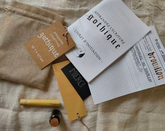 May include: A set of calligraphy supplies for learning gothic script. The set includes a booklet titled "Apprendre l'écriture gothique - Livret d'initiation", a small burlap bag, a brown paper tag with the text "Apprendre l'écriture gothique de 7 à 107 ans", a black card with the word "Merci" written in white, and a small bottle of ink.