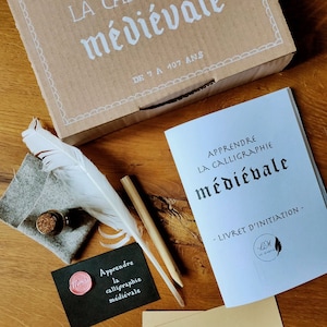 Puede incluir: Un kit de caligrafía con un libro titulado "Apprendre la calligraphie médiévale - Livret d'initiation", una pluma, un tintero y un sello de cera. El kit está sobre una superficie de madera.