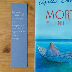 May include: A rectangular periwinkle bookmark featuring white hand-lettered French text that reads: La vraie amiti&eacute; ce n'est pas d'&ecirc;tre ins&eacute;parable, c'est d'&ecirc;tre s&eacute;par&eacute; et que rien ne change. Beside it lies a book titled Mort sur le Nil.
