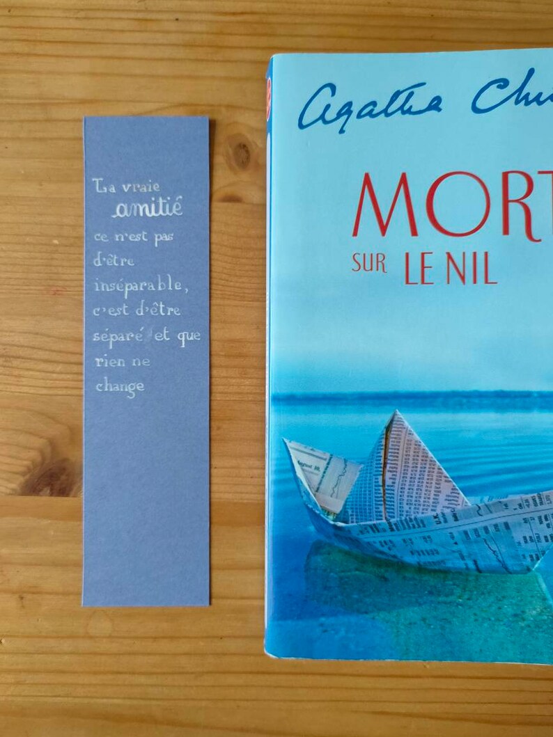 May include: A rectangular periwinkle bookmark featuring white hand-lettered French text that reads: La vraie amiti&eacute; ce n'est pas d'&ecirc;tre ins&eacute;parable, c'est d'&ecirc;tre s&eacute;par&eacute; et que rien ne change. Beside it lies a book titled Mort sur le Nil.