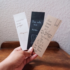 May include: Three rectangular paper bookmarks. The white one features the text encore une page and a book illustration. The black one displays Keep calm and READ with a scroll. The brown one lists the word read in various languages and a bookmark icon.