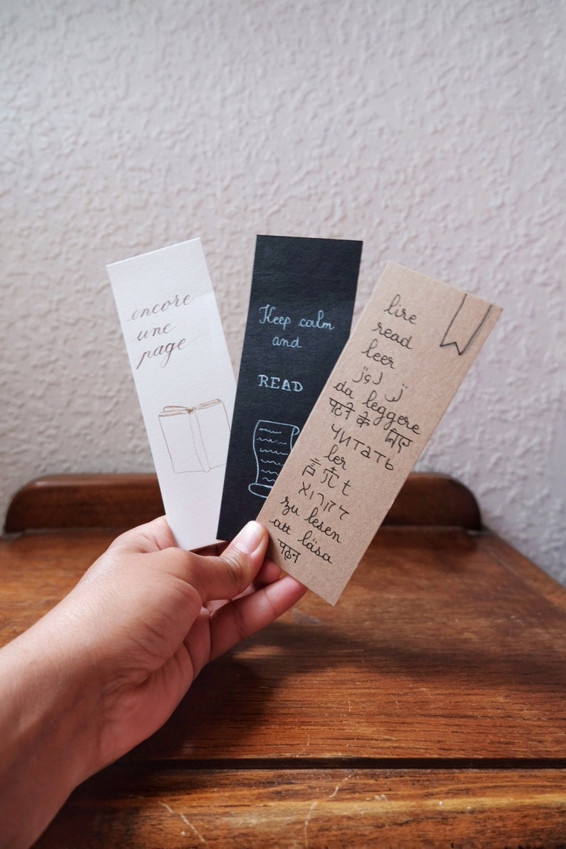 May include: Three rectangular paper bookmarks. The white one features the text encore une page and a book illustration. The black one displays Keep calm and READ with a scroll. The brown one lists the word read in various languages and a bookmark icon.