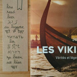 May include: A rectangular tan paper bookmark featuring the word read in various languages and a hand-drawn bookmark icon, placed next to a book titled LES VIKINGS with a cover illustration of a wooden Viking ship prow on the water.