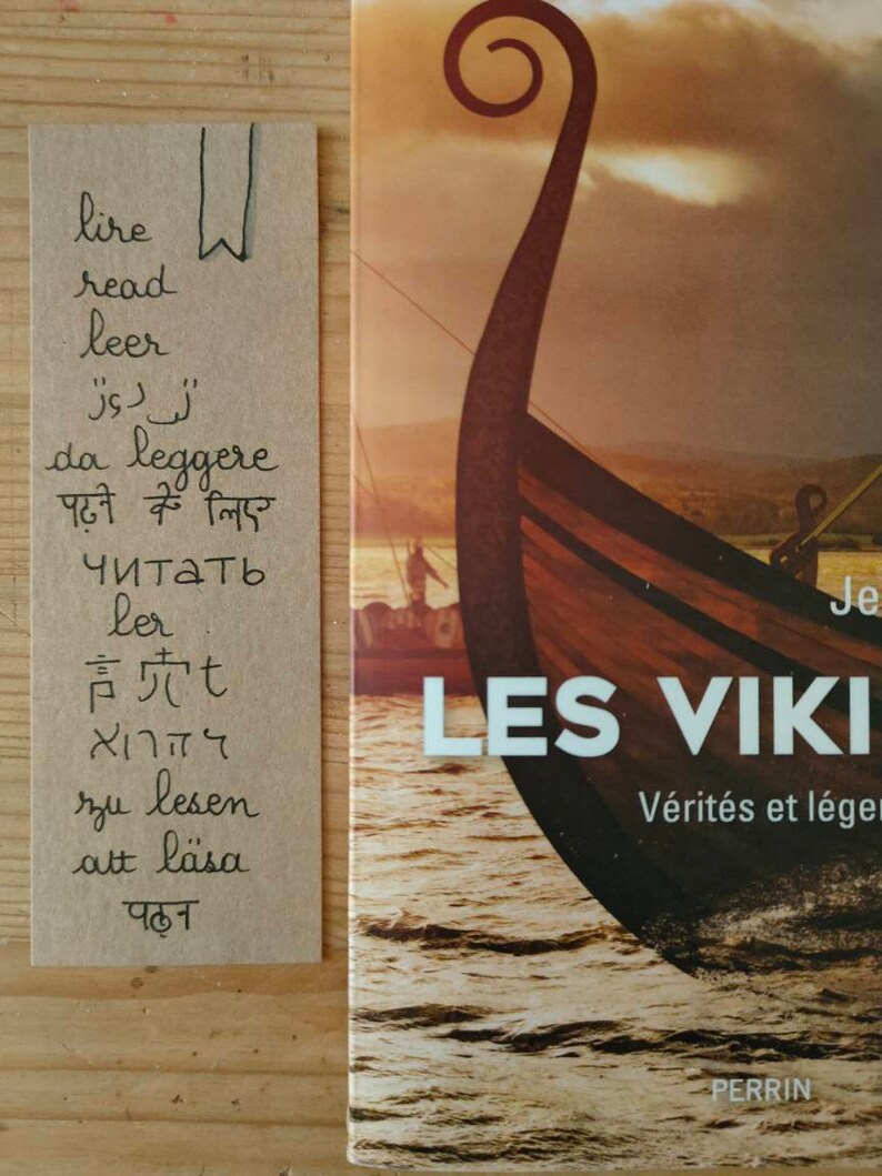 May include: A rectangular tan paper bookmark featuring the word read in various languages and a hand-drawn bookmark icon, placed next to a book titled LES VIKINGS with a cover illustration of a wooden Viking ship prow on the water.