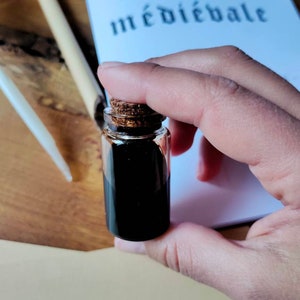 May include: A small glass bottle with a cork stopper filled with black ink. The bottle is being held by a hand. The bottle is sitting on a white surface with the text "Apprendre la calligraphie m&eacute;di&eacute;vale" visible in the background.