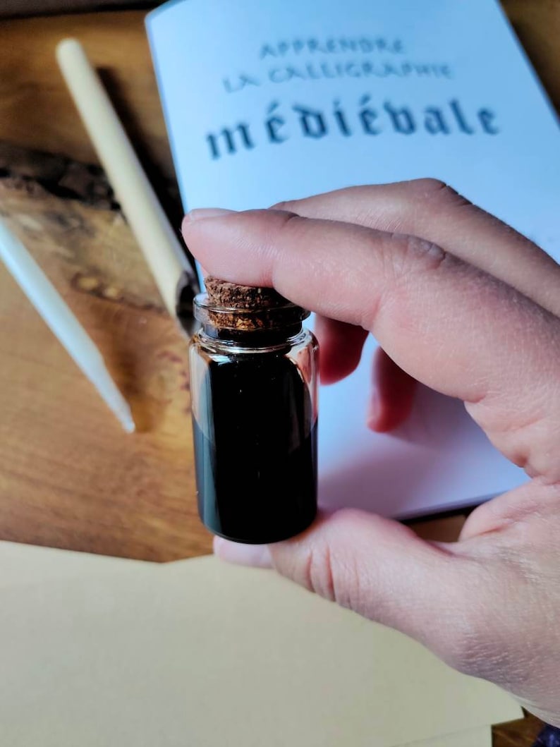 May include: A small glass bottle with a cork stopper filled with black ink. The bottle is being held by a hand. The bottle is sitting on a white surface with the text "Apprendre la calligraphie m&eacute;di&eacute;vale" visible in the background.