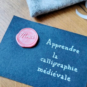 May include: A black card with white text that reads "Apprendre la calligraphie m&eacute;di&eacute;vale". A pink wax seal with the word "Merci" is on the card.