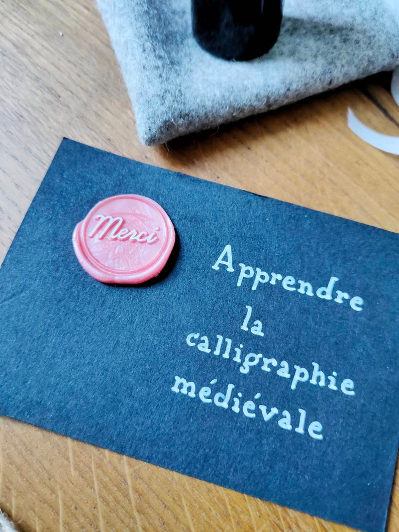 May include: A black card with white text that reads "Apprendre la calligraphie m&eacute;di&eacute;vale". A pink wax seal with the word "Merci" is on the card.