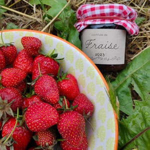 Puede incluir: Un cuenco lleno de fresas rojas maduras, con tallos y hojas verdes. En el fondo, un tarro de mermelada de fresa con tapa a cuadros rojos y blancos. La etiqueta del tarro dice "confiture de Fraise 2023".