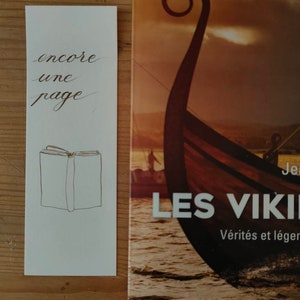May include: A white rectangular bookmark featuring the black cursive text encore une page and a simple line drawing of an open book, positioned next to a book cover titled Les Vikings with an illustration of a wooden ship on the water.