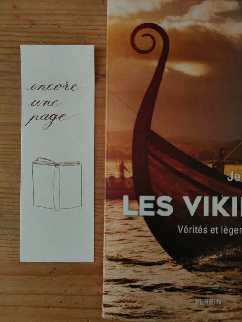 May include: A white rectangular bookmark featuring the black cursive text encore une page and a simple line drawing of an open book, positioned next to a book cover titled Les Vikings with an illustration of a wooden ship on the water.