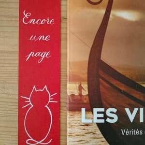 May include: A rectangular red bookmark featuring the white handwritten text Encore une page and a white line drawing of a cat silhouette at the bottom, positioned next to a book cover showing a Viking ship prow on water at sunset.