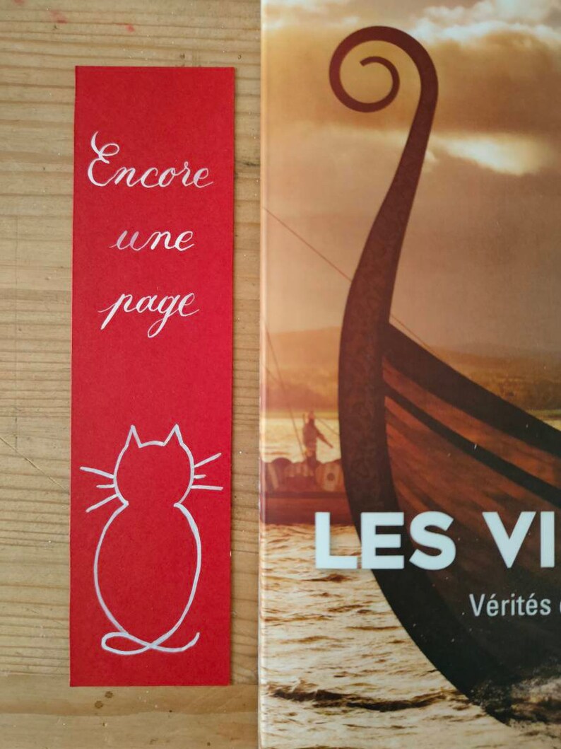 May include: A rectangular red bookmark featuring the white handwritten text Encore une page and a white line drawing of a cat silhouette at the bottom, positioned next to a book cover showing a Viking ship prow on water at sunset.