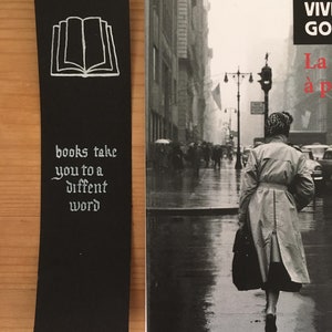 May include: A black rectangular bookmark featuring a white line drawing of an open book at the top and the white text "books take you to a diffent word" below it, positioned next to a book cover showing a black and white street scene.