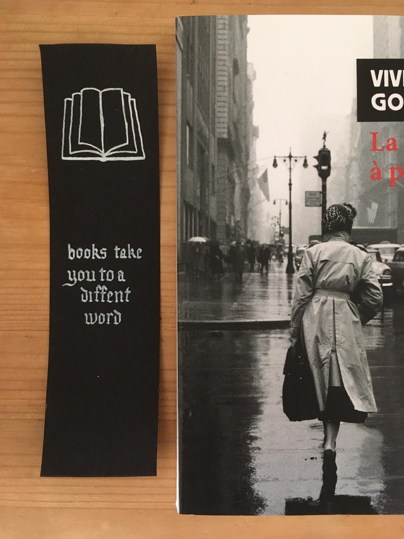 May include: A black rectangular bookmark featuring a white line drawing of an open book at the top and the white text "books take you to a diffent word" below it, positioned next to a book cover showing a black and white street scene.