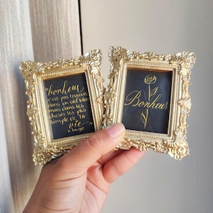 Peut inclure: Deux petits cadres photo rectangulaires avec des bordures métalliques dorées ornées. Chaque cadre contient un fond noir avec une calligraphie dorée. L'un affiche une rose et le mot Bonheur, l'autre présente un texte en français.