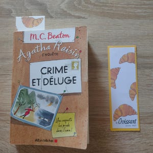 May include: A paperback book titled Agatha Raisin Crime et D&eacute;luge by M.C. Beaton with a tan cover featuring a storm illustration and a yellow sticky note, accompanied by a rectangular bookmark decorated with a repeating croissant pattern.