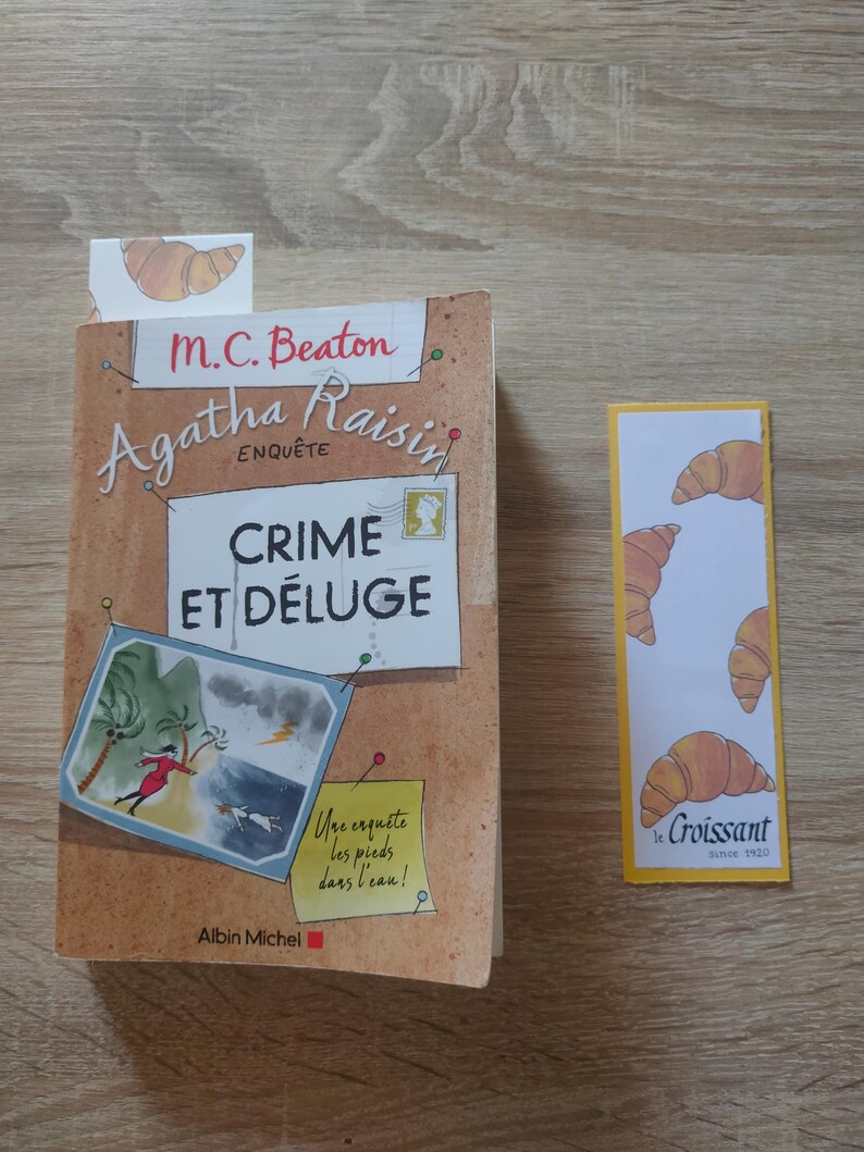 May include: A paperback book titled Agatha Raisin Crime et D&eacute;luge by M.C. Beaton with a tan cover featuring a storm illustration and a yellow sticky note, accompanied by a rectangular bookmark decorated with a repeating croissant pattern.