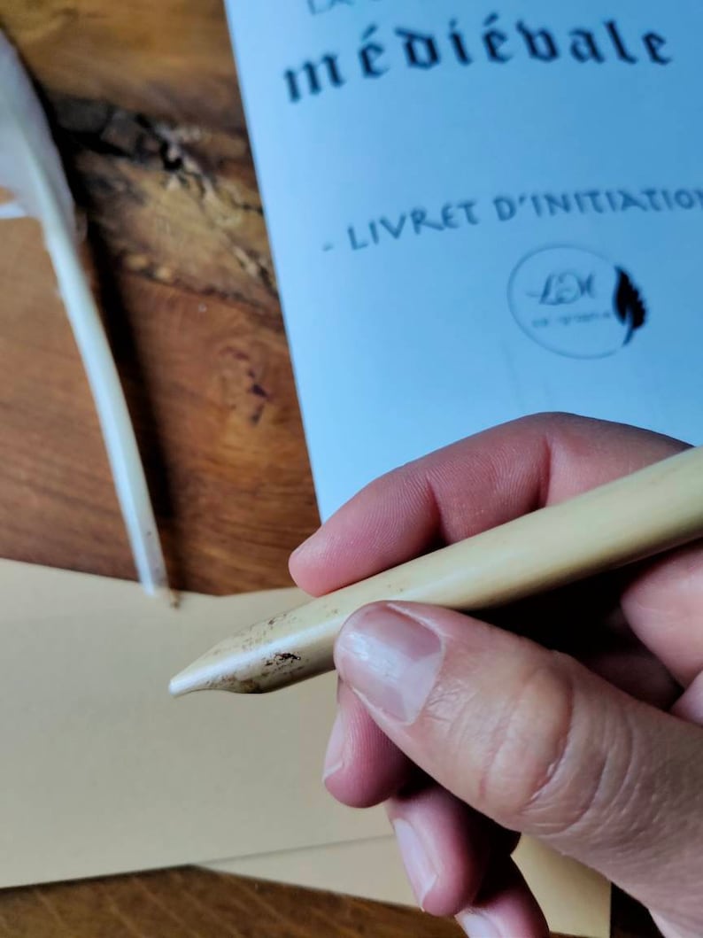 May include: A close-up of a person's hand holding a light brown calligraphy pen with a pointed tip. The pen is resting on a beige piece of paper. The background is a wooden surface.