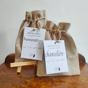 Peut inclure: Deux sacs en toile de jute avec des étiquettes blanches. Les étiquettes indiquent "APPRENDRE L'ECRITURE humanistique DE 7 A 107 ANS" et "APPRENDRE L'ECRITURE chancelière DE 7 A 107 ANS". Les sacs sont noués avec de la ficelle et posés sur une surface en bois.