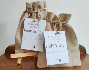 May include: Two burlap drawstring bags with white tags. The tags read "APPRENDRE L'ECRITURE humanistique DE 7 A 107 ANS" and "APPRENDRE L'ECRITURE chancelière DE 7 A 107 ANS". The bags are tied with twine and sit on a wooden surface.