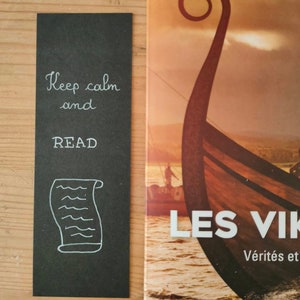May include: A rectangular black bookmark featuring white hand-lettered text that reads Keep calm and READ, accompanied by a white line drawing of a scroll. The bookmark is positioned next to a book cover depicting a wooden Viking ship on water.