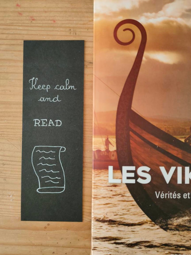 May include: A rectangular black bookmark featuring white hand-lettered text that reads Keep calm and READ, accompanied by a white line drawing of a scroll. The bookmark is positioned next to a book cover depicting a wooden Viking ship on water.