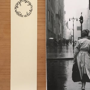May include: A cream-colored rectangular bookmark featuring a circular design made of the repeated word "lecture" in black script, positioned next to a black and white photograph of a person in a trench coat walking down a rainy city street.